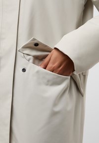 Manteau beige clair avec une texture lisse et mate. Le design comprend une poche latérale avec un bouton noir et une coupe décontractée.