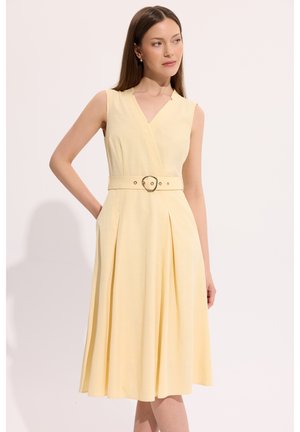 Kobieta w bladożółtej, bezrękawnej sukience midi z paskiem w talii i plisowaną spódnicą, stojąca z jedną ręką w kieszeni.