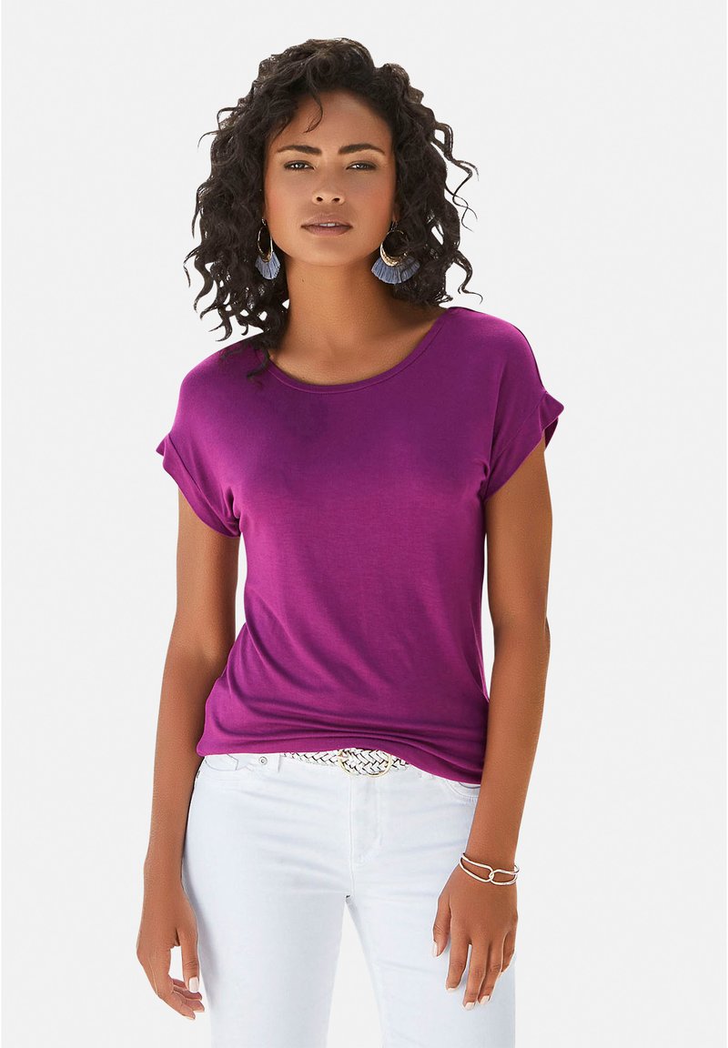 Lila Kurzarm-T-Shirt aus einem glatten, dehnbaren Stoff mit umgeschlagenen Ärmeln und rundem Ausschnitt, kombiniert mit weißen Hosen und einem dekorativen Gürtel.