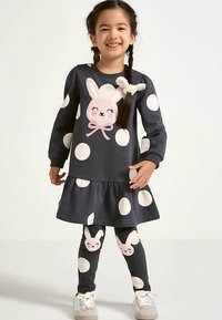 Harmaa mekko, jossa suuria valkoisia pallokuvioita, röyhelöreuna ja vaaleanpunainen pupuapplikaatio. Yhteensopivat leggingsit, joissa pupukuviot, yhdistettynä tennareihin.