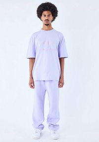 T-shirt oversized lilla con logo rosa e pantaloni sportivi abbinati. Tessuto morbido simile al cotone, vestibilità rilassata e sneaker bianche completano il look.