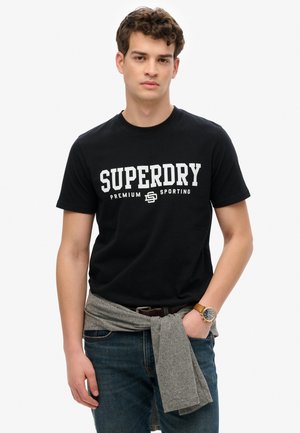 Zwarte katoenen t-shirt met witte tekst "SUPERDRY PREMIUM SPORTING". Gecombineerd met blauwe jeans, een grijze trui om de taille geknoopt, en een horloge.