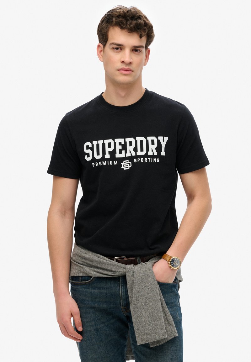 Sort bomulds t-shirt med hvid tekst "SUPERDRY PREMIUM SPORTING". Sammen med blå jeans, en grå sweater bundet om taljen og et ur.