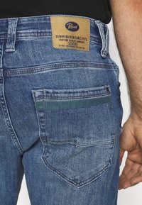 Kék farmernadrág, egyenes szabással, bőr címkével a hátsó derékrészen és zöld akcentussal a hátsó zsebnél.