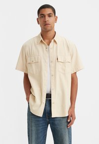 Short-sleeve button-up shirt in light beige fabric, featuring two front pockets, a relaxed fit, and silver hardware details. Worn with blue jeans.
