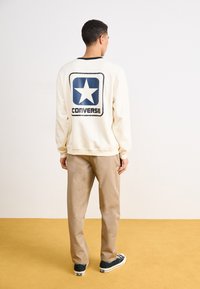 Krémová mikina s modrým logom hviezdy a nápisom "CONVERSE"; kombinovaná s béžovými nohavicami a čiernymi teniskami na žltom podlahe.