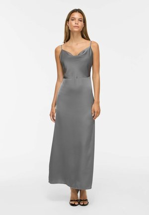 Femme portant une robe maxi en satin gris sans manches avec des bretelles fines et des talons ouverts noirs, debout devant un fond blanc uni.