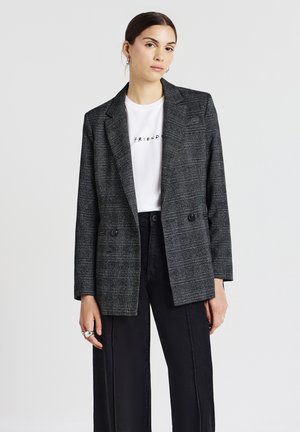 Blazer grigio a quadri con vestibilità sartoriale, due bottoni frontali e rever a punta, indossato sopra una t-shirt bianca e pantaloni a gamba larga neri.