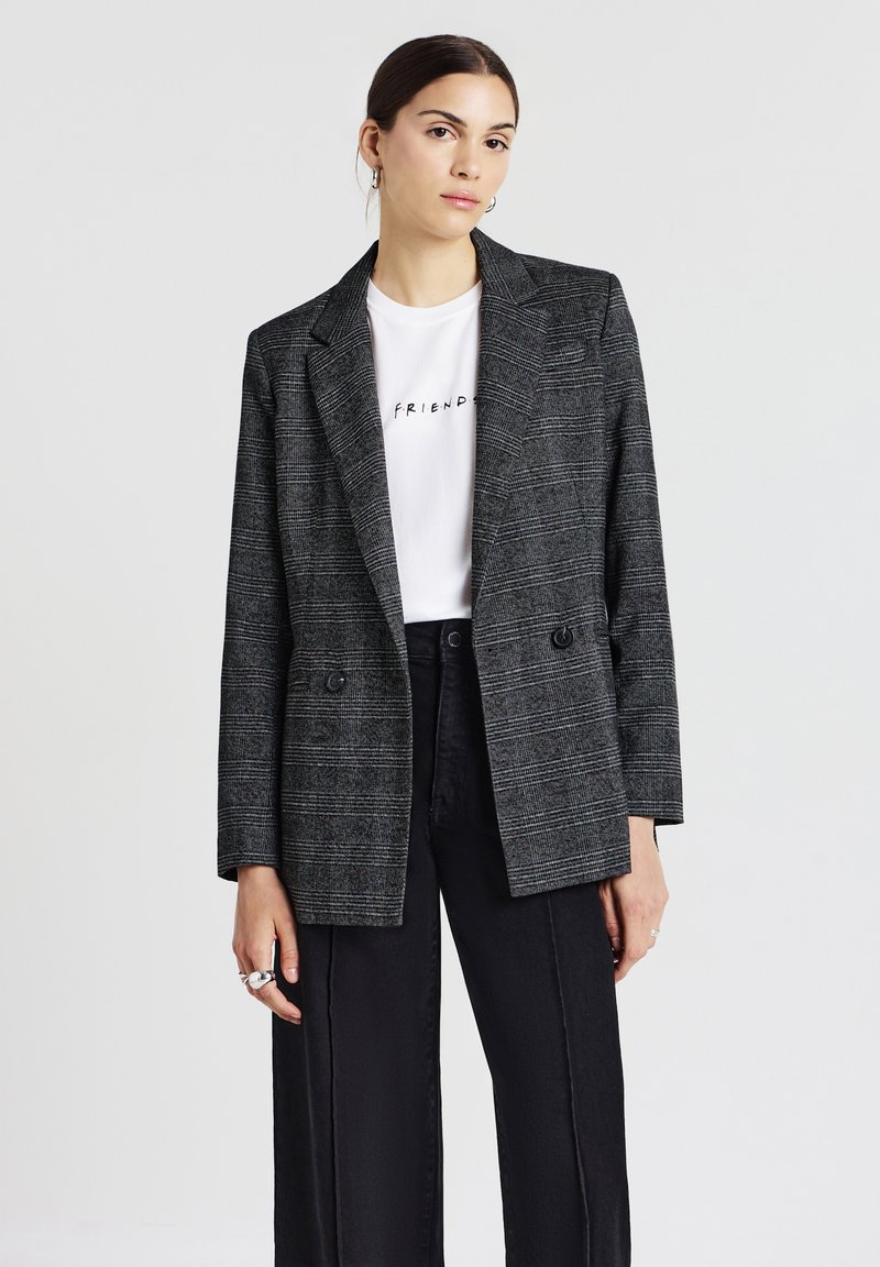 Blazer grigio a quadri con vestibilità sartoriale, due bottoni frontali e rever a punta, indossato sopra una t-shirt bianca e pantaloni a gamba larga neri.