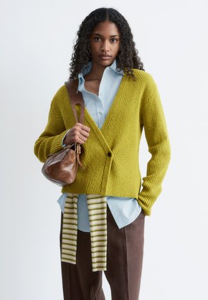 Kobieta z kręconymi włosami, ubrana w jasnoniebieską koszulę, kardigan z dzianiny w kolorze musztardowym, brązowe spodnie i trzymająca brązową skórzaną torbę na ramię.