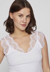 Witte tanktop met ingewikkelde witte kanten rand op de schouders. De stof lijkt geribbeld te zijn, met een diepe V-hals en een gladde textuur.