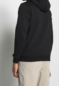 Černá mikina s texturovanou látkou a nastavitelnou kapucí na šňůrku; žebrované manžety a spodní lem. Spárovaná s světlými kalhotami.