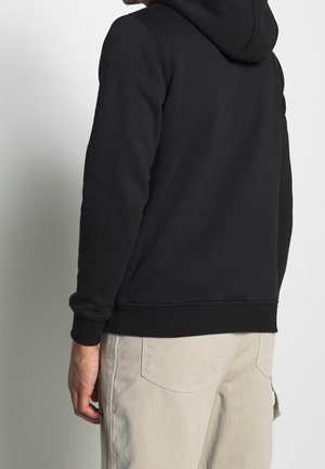 Hoodie noir en tissu texturé avec capuche à cordon de serrage ajustable ; poignets et ourlet inférieur côtelés. Accordé avec un pantalon de couleur claire.