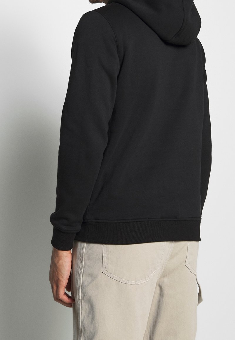 Černá mikina s texturovanou látkou a nastavitelnou kapucí na šňůrku; žebrované manžety a spodní lem. Spárovaná s světlými kalhotami.