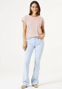 Gestreiftes Kurzarmoberteil in Weiß und Rot, kombiniert mit hellblauen Schlagjeans. Das Model steht mit einer Hand in der Tasche und trägt goldene Schuhe.