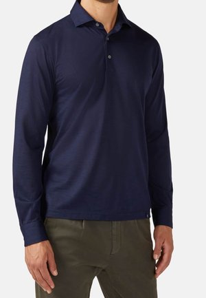 Mann, der ein marineblaues Poloshirt mit langen Ärmeln und olivgrüne Hose trägt, steht mit entspannt an den Seiten hängenden Armen.