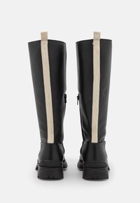 Une paire de bottes noires montant jusqu'aux genoux avec des tirettes verticales beiges et des semelles épaisses, vues de l'arrière sur un fond blanc.