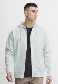 Lyseblå hettejakke med glidelås i myk stoff, med snøring på hetten, frontlommer og en avslappet passform. Brukt over en svart skjorte.
