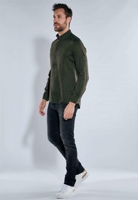 Olivgrüne, knöpfbare Hemd aus strukturiertem Stoff, kombiniert mit dunklen Slim-Fit-Jeans und weißen Sneakers mit hellen Akzenten.