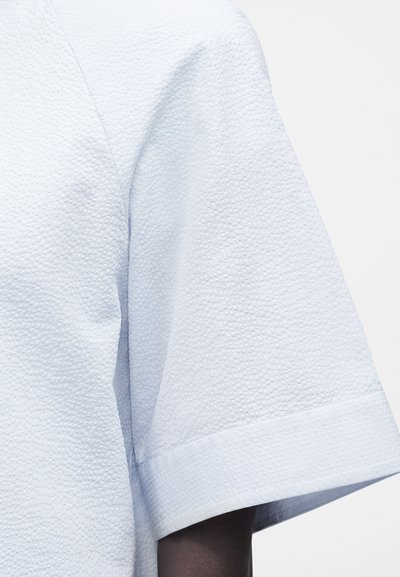 Κοντινό πλάνο ενός γαλάζιου υφασμάτινου πουκαμίσου με κοντά μανίκια σε άτομο με σκουρόχρωμο δέρμα, εστιάζοντας στο ύφασμα και τη λεπτομέρεια του μανικιού.