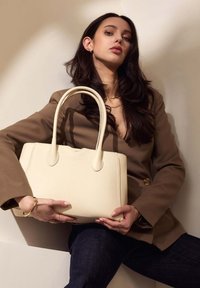 Sac à main en cuir couleur crème avec une forme structurée, deux poignées et une texture lisse, tenu dans les mains contre un fond neutre.
