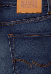 Vaqueros de mezclilla azul oscuro con costuras naranjas, que presentan una etiqueta de cuero rectangular con la inscripción "JACK & JONES" en la cintura.
