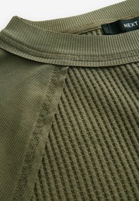 Haut vert olive à texture côtelée, avec un col lisse et des détails de coutures contrastantes. Étiquette visible à l'intérieur du col.