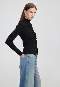 Czarna bluzka z długim rękawem i wysokim kołnierzem, zdobiona pionowymi falbanami oraz guzikami z przodu, zestawiona z jasnoniebieskimi spodniami typu wide-leg.