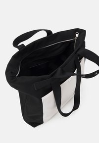 Borsa tote in tela nera con un pannello accentato bianco; presenta una chiusura superiore con zip, due manici robusti e una forma non strutturata.