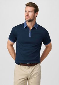 Man in een donkerblauw poloshirt met korte mouwen en lichtblauwe bies, en beige broek, staand met handen achter de rug tegen een witte achtergrond.