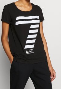 Černé tričko s velkým bílým grafickým motivem z flitrů v pruhovaném designu. Měkká látka, kulatý výstřih, s logem "EA7 Emporio Armani".
