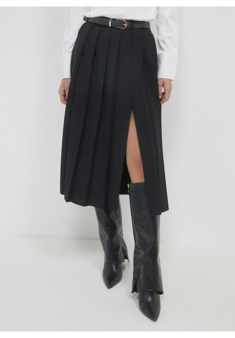 Jupe midi plissée noire avec une fente frontale, associée à des bottes en cuir noires montantes à bout pointu et un haut blanc à manches longues.