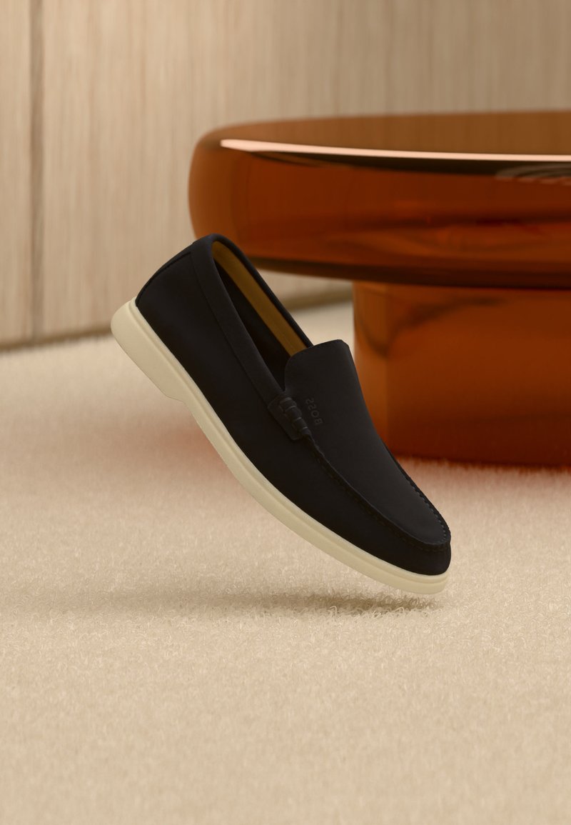 Mocassino nero slip-on con suola bianca poggiato su moquette beige accanto a una base di tavolo rotonda di colore ambra.