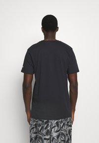 Muž otočený zády, oblečený v jednobarevném černém tričku a šedých šortkách s černými abstraktními vzory štětcových tahů na bílém pozadí.