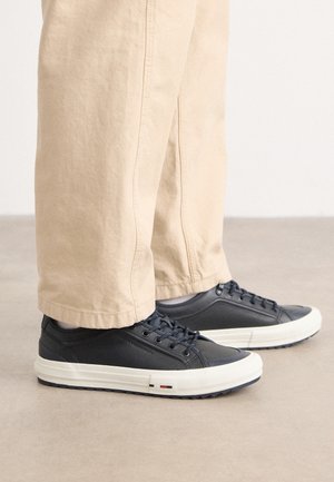 Persona con pantalones beige y zapatillas negras bajas con suelas blancas, de pie sobre un suelo gris liso.