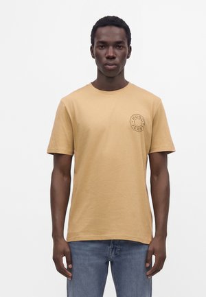 Jeune homme portant un t-shirt Hugo Jeans marron clair et un jean bleu, debout devant un fond blanc uni.