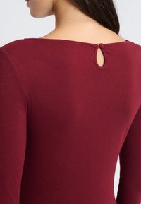 Burgunderrotes Langarmshirt mit rundem Ausschnitt und einem kleinen Schlitzdetail hinten, aus weichem, glattem Stoff gefertigt. Schlichtes, enganliegendes Design.