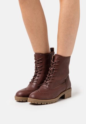 Botines de cuero marrón con detalles brogue, cremallera lateral, cordones y tacón bajo apilado, usados en piernas desnudas.