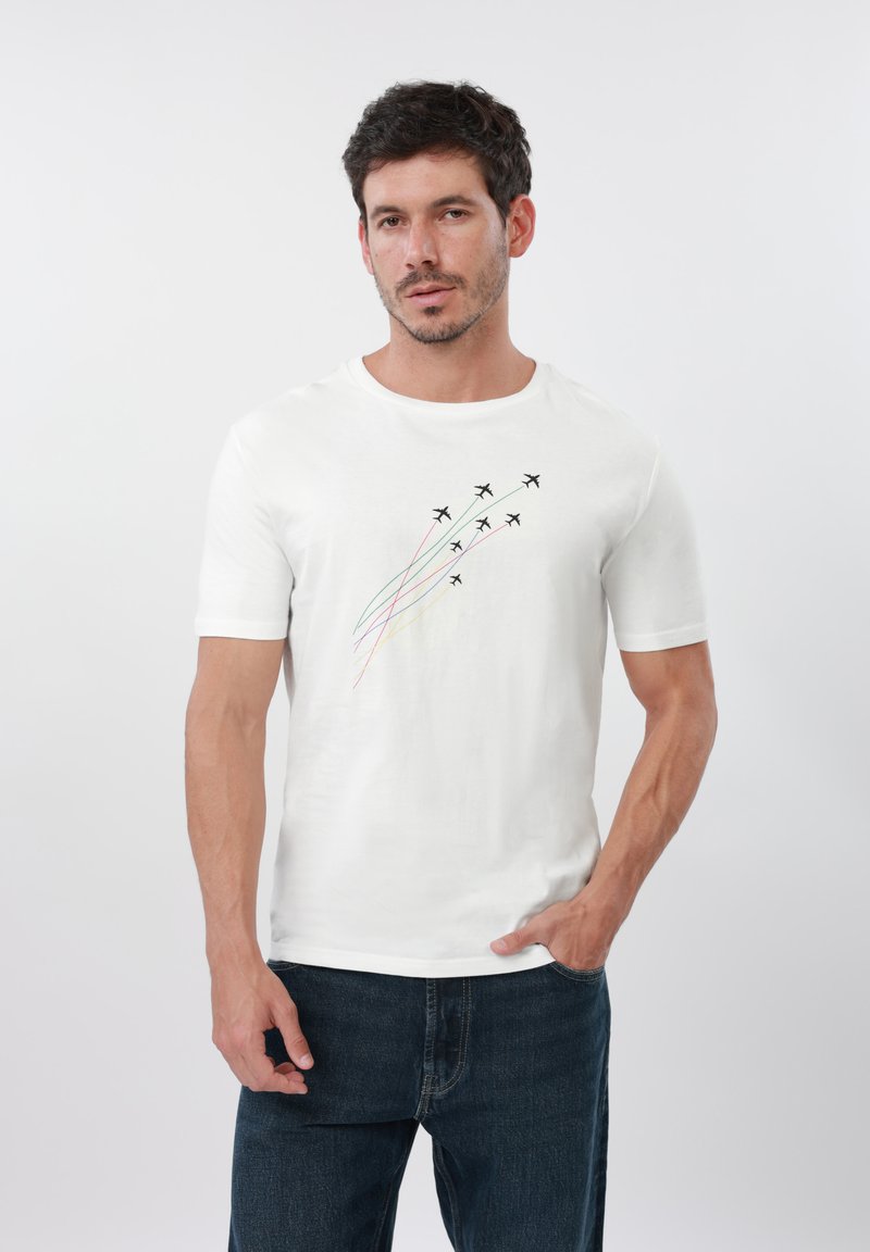 Άνδρας που φοράει λευκό μπλουζάκι με επτά μικρά μαύρα αεροπλάνα να πετούν διαγώνια προς τα πάνω, το καθένα με έγχρωμη ουρά, συνδυασμένο με μπλε τζιν.