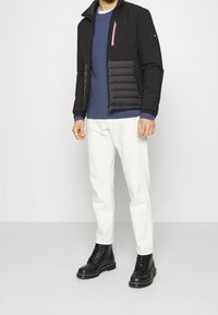 Schwarze Herrenjacke mit gepolstertem Einsatz, dunkelblauer strukturiertem Pullover, weißen Hosen und schwarzen Knöchelschuhen. Mit roten und weißen Reißverschlussakzenten.