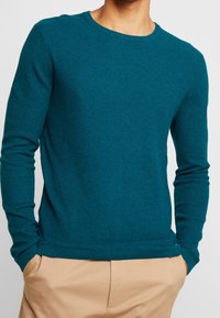 Man i en teal-färgad texturerad långärmad tröja som är något instoppa i beiga byxor, med händerna i fickorna, mot en vit bakgrund.