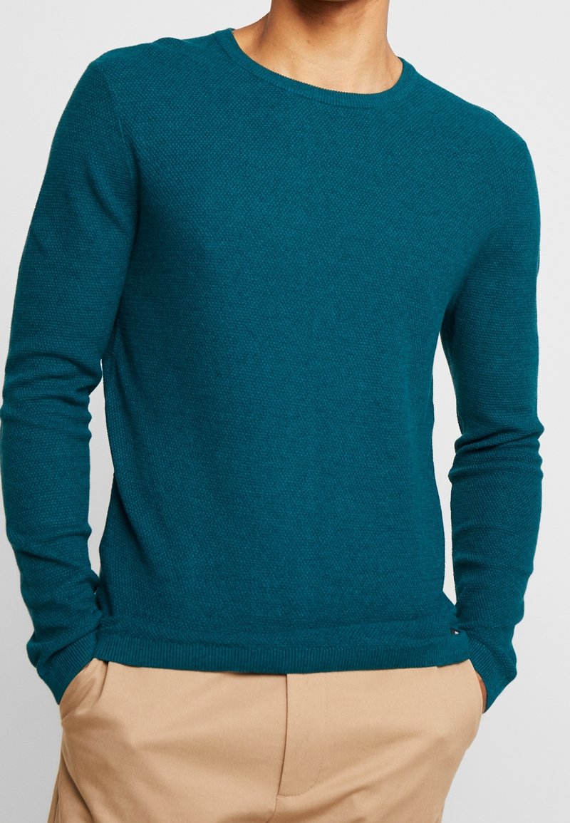 Man i en teal-färgad texturerad långärmad tröja som är något instoppa i beiga byxor, med händerna i fickorna, mot en vit bakgrund.