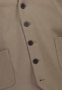Braunes Jackenmaterial mit texturierter Oberfläche, ausgestattet mit sechs schwarzen Knöpfen auf der Vorderseite und einer einzigen Tasche auf der linken Seite.
