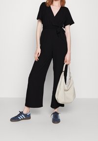 Mono negro con mangas cortas, escote en V profundo, cintura atada y silueta de pierna ancha. Combinado con zapatillas deportivas azul marino y un bolso beige claro.