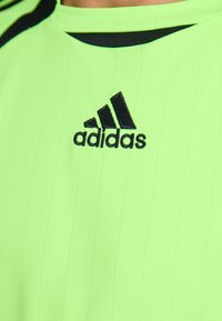 Žalias marškinėlis su juodais akcentais ir vertikaliomis juostelėmis. Viršutinėje krūtinės dalyje yra juodas siuvinėtas Adidas logotipas.