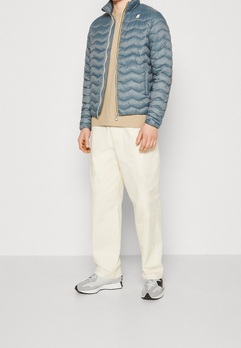 Mann in einer hellblauen Steppjacke, beigem Hemd, cremefarbenen Hosen und grauen Sneakers, der vor einem einfarbigen hellen Hintergrund steht.