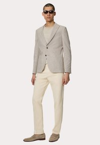 Lys grå linned blazer med to knapper, cremefarvet t-shirt, cremefarvede bukser og grå loafers. Modellen bærer mørke solbriller.