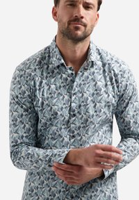 Man in een langärmige, knoopsluitende blouse met een abstract blauw en grijs patroon, terwijl hij zijn linkerschoudermanchet aanpast.