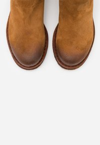 Stivaletti in suede marrone con punta arrotondata, caratterizzati da una cucitura marrone a contrasto lungo la suola e una texture liscia.