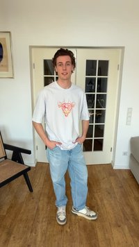 Jeune homme portant un t-shirt blanc avec un motif graphique, un jean bleu clair et des baskets, debout à l'intérieur avec les mains dans les poches, sol en bois et porte à panneaux de verre derrière lui.